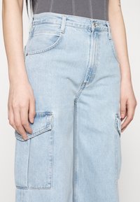 Svetle modré džíny s vysokým pasem, cargo styl, se dvěma bočními kapsami a volným střihem. Materiál vykazuje mírné vyblednutí a strukturu.