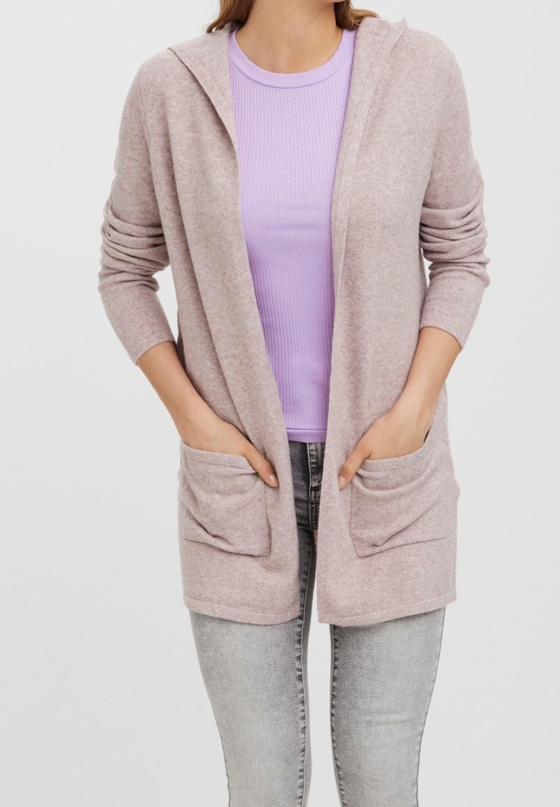 Lichtroze cardigan met een zachte textuur en een capuchon, een open voorkant en twee steekzakken aan de voorkant, gedragen over een lila top met korte mouwen en grijze spijkerbroek.