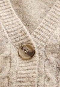 Beżowy kardigan wykonany na drutach, z teksturowanym wzorem, ściągaczem przy kołnierzu oraz błyszczącym, okrągłym guzikiem z dwoma otworami z przodu.