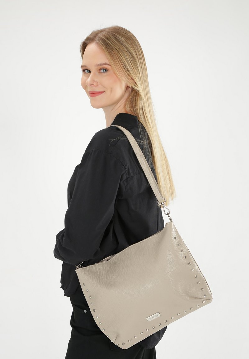 Beige leren handtas met metalen studs langs de rand, uitgerust met een afneembare schouderband en een gladde textuur.