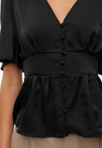 Svart peplumbluse laget av satengstoff, med dyp V-utringning, korte ermer og midtstilte knapper med myke runde detaljer.