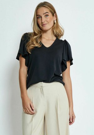 Blouse noire à col en V avec des manches courtes bouffantes, en tissu lisse. Associée à un pantalon beige taille haute, créant un look contrasté.