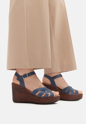Sandalias cuña azules con correas de cuero cruzadas, suela de madera texturizada y hebilla en el tobillo. Combinadas con pantalones beige claros de pierna ancha.