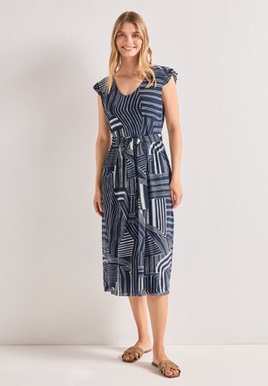Jeune femme portant une robe midi bleu marine à motifs géométriques, manches courtes à volants et sandales beiges à enfiler.