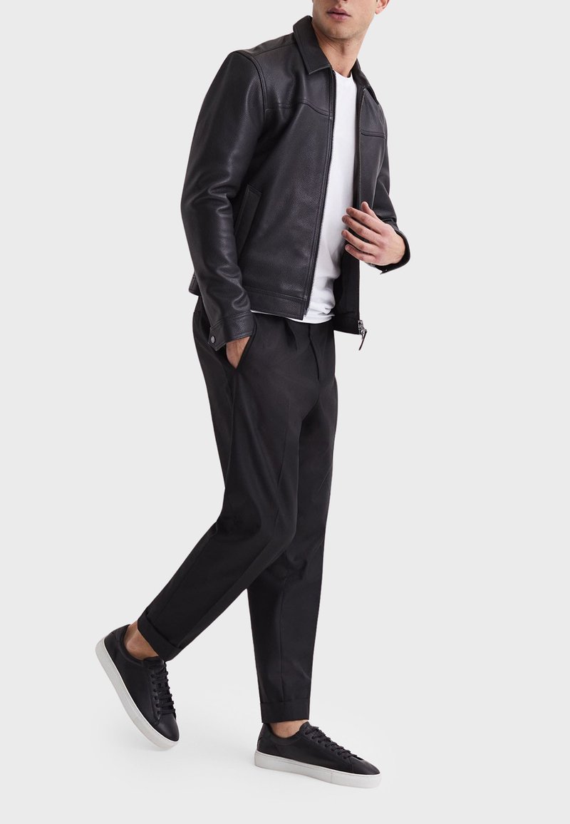 Mann trägt schwarze Lederjacke, weißes T-Shirt, schwarze Hose und schwarze Sneaker mit weißen Sohlen vor einfarbigem Hintergrund.