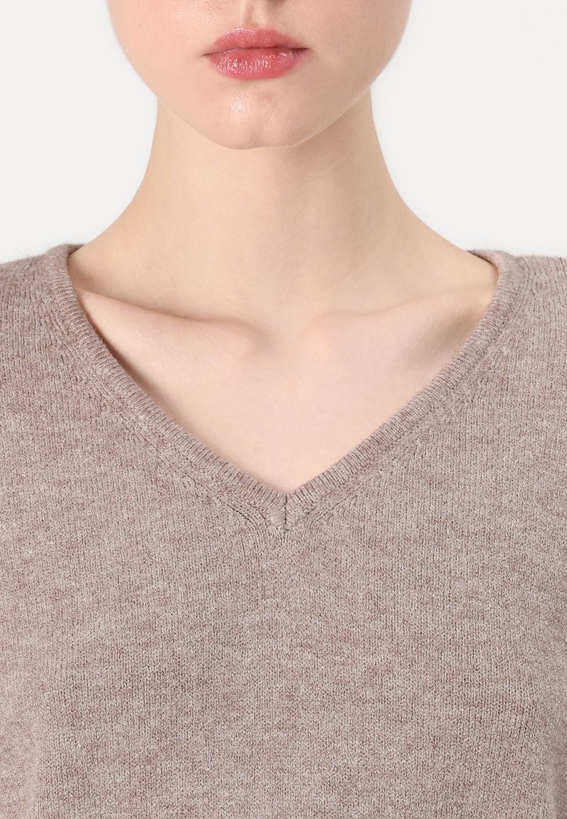Pull en V en tissu tricoté beige doux. Présente une texture lisse avec des détails côtelés le long de l'encolure, mettant en valeur la forme du vêtement.