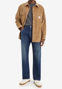 Chaqueta de camisa de pana marrón con un bolsillo en el pecho, combinada con vaqueros azules de pierna recta. Zapatillas blancas con suelas marrón claro completan el conjunto.