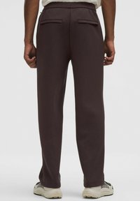 Dunkelbraune Hose mit glatter Textur, elastischem Bund und zwei Gesäßtaschen. Kombiniert mit hellen Sneakern.