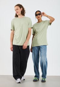 Light green cotton t-shirts, relaxed fit. The male model wears black wide-leg jeans and white sneakers, while the female has blue jeans and dark shoes.