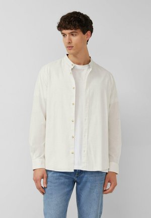 Jeune homme aux cheveux courts et bouclés portant une chemise blanche à col mandarin boutonnée sur un t-shirt blanc et un jean bleu clair, regardant vers la gauche.