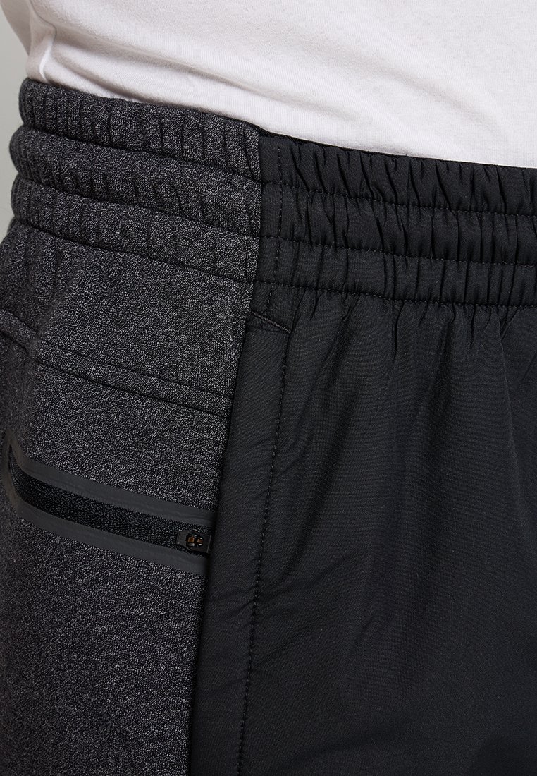 Primer plano de pantalones con cinturilla elástica bicolor, fabricados en tela texturizada gris oscuro y tela negra lisa, que cuentan con un bolsillo con cremallera en el lateral.