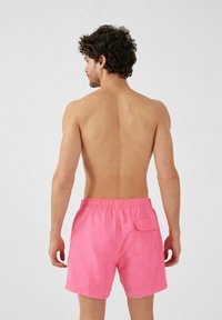 Costume da bagno rosa con vita elastica e tasca laterale, realizzato in tessuto leggero. Vista da dietro, che mostra un schiena e spalle muscolose.