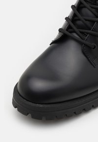 Bota de tobillo de cuero negro con punta redondeada, superficie texturizada, suela gruesa negra y cordones negros, con ojales de metal para la inserción del cordón.
