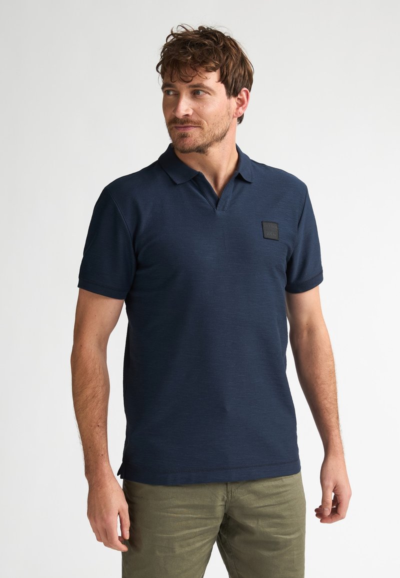 Polo bleu marine à manches courtes, tissu texturé et écusson sur la poitrine. Coupe ajustée avec un col classique et une fente latérale pour faciliter les mouvements.