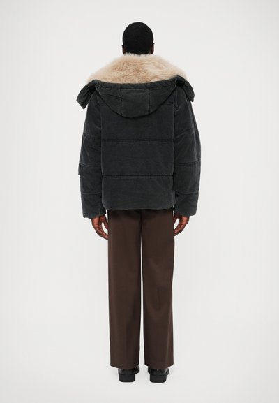 Sort pufferjakke med en tan faux fur hætte, der har en firkantet form og vandrette kviltede linjer, parret med brune bukser og sko.