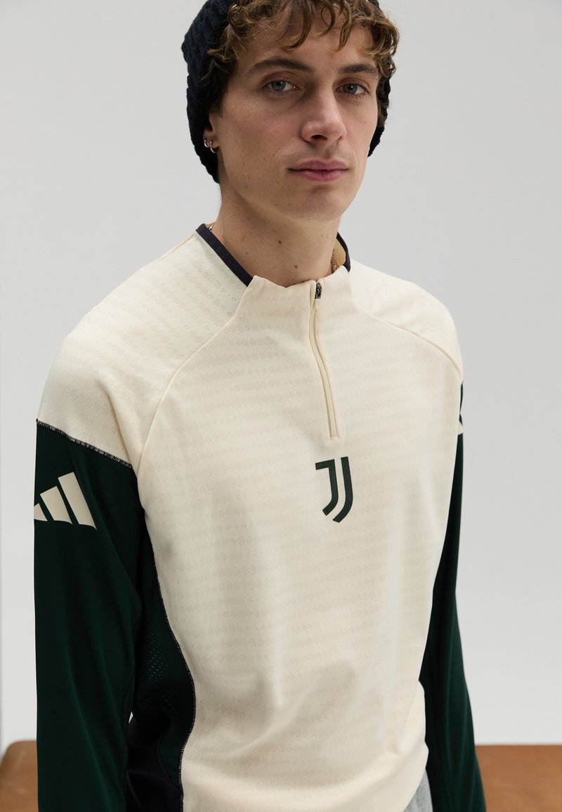Cream and green zip-up sports top, featuring a geometric pattern, a high collar, and a logo on the chest, made of textured fabric.