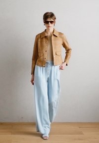 Svetlehnědá semišová bunda s předními knoflíky a kapsami, spárovaná se světle modrými kalhotami s širokými nohavicemi a sandály. Model má na sobě sluneční brýle a stojí na dřevěné podlaze.