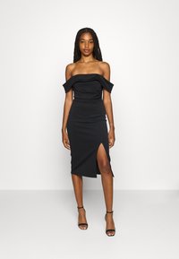 Robe noire décolletée en épaules, fabriquée en tissu extensible, avec une silhouette ajustée et une fente haute sur le côté gauche.