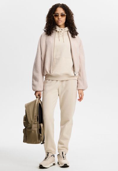Woman with curly hair wearing beige hoodie, light pink jacket, beige pants, and sneakers holding a beige backpack against white background.