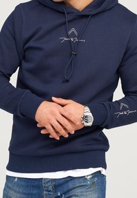 Marinblå huvtröja i bomullsblandning, med en framficka, dragsko på huvan och logotyp på vänster ärm. Bärs över en vit T-shirt.