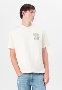 Cream-colored t-shirt featuring a floral design in green and yellow with "GUESS DESIGN" text. Made of soft cotton with a relaxed fit.