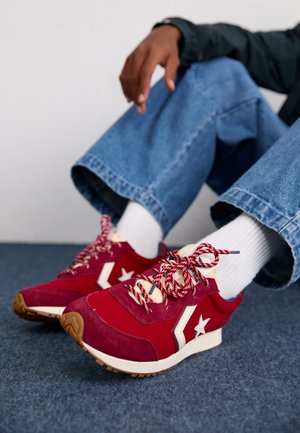 Personne portant un jean bleu et des chaussettes blanches, assise jambes croisées, portant des baskets rouges avec une étoile blanche et des lacets à motifs.