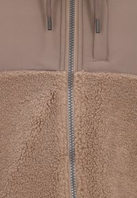 Braune Jacke mit glattem Oberstoff und strukturiertem Fleece im unteren Bereich. Ein silberner Reißverschluss verläuft in der Mitte, mit verstellbaren Kordelzügen.