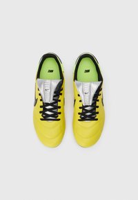 Zapatillas deportivas amarillas con cordones negros, detalles plateados y una superficie texturizada. El interior cuenta con una plantilla verde brillante.
