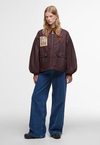 Μπορντό bomber jacket με λουλούδι, φερμουάρ μπροστά, δύο μπροστινές τσέπες και καφέ δερμάτινες λεπτομέρειες. Συνδυάζεται με μπλε παντελόνι φαρδύ.