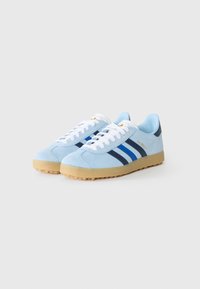 Lyseblå ruskinnsneakers med hvite snøringer, med tre mørkeblå striper, en beige såle, og "Gazelle"-tekst i gull på siden.