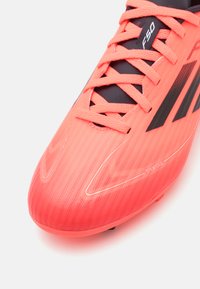 Botas de fútbol de un brillante color coral, con un diseño aerodinámico y elegante, con acentos negros, superficie texturizada y un sistema de cordones con un patrón.
