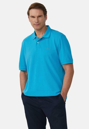 Türkises Polo-Shirt mit kurzen Ärmeln, kragenähnlichem Ausschnitt und subtiler Texturdetails; getragen mit dunkelblauen Hosen. Besticktes Logo auf der Brust.