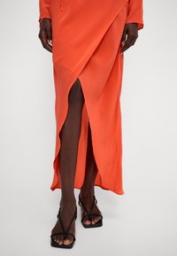 Langes oranges Kleid aus weichem Stoff, mit einem tiefen Schlitz und einer lockeren Passform. Getragen mit schwarzen Sandalen mit Riemen, die ein minimalistisches Design zeigen.