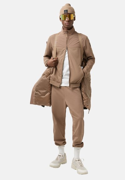 Tan tracksuit with a zip-up jacket and tapered pants, complemented by a white shirt, a beanie, and reflective sunglasses. Chunky sneakers.