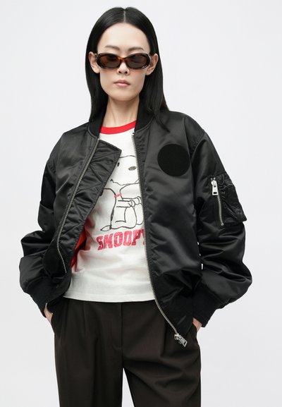 Crna saten bomber jakna s okruglim flasterom, rebrastim rukavima i džepom na zatvarač. Nosila se preko bijele majice s grafikom sa Snoopyjem. Smeđe hlače.
