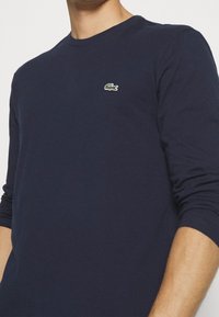 Granatowa koszulka z długim rękawem wykonana z bawełny, z małym haftowanym logo krokodyla w kolorze zielonym i fioletowym na lewej piersi.