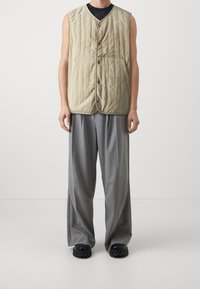 Beżowa pikowana kamizelka z dekoltem w serek, zapięciem na guziki z przodu oraz subtelnym logo, połączona z szarymi spodniami o szerokich nogawkach i czarnym, masywnym obuwiem.