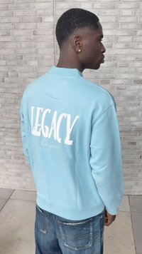 Mladý muž ve světle modré mikině s textem "LEGACY Original" na zadní straně, stojící bočně proti šedé cihlové zdi.