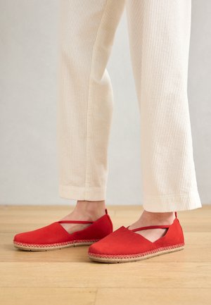 Κόκκινα εσπαντρίγια με σουέτ άνω μέρος, κλειστή μύτη και διασταυρούμενες λωρίδες. Φυσική υφαντή βάση με πλεχτή υφή. Ζευγάρι που φοριέται με κρεμ παντελόνια από κορντούρα.