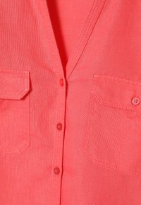 Camicia di lino corallo con scollatura a V, due tasche sul petto e bottoni rossi. Texture liscia con dettagli di cucitura a vista.