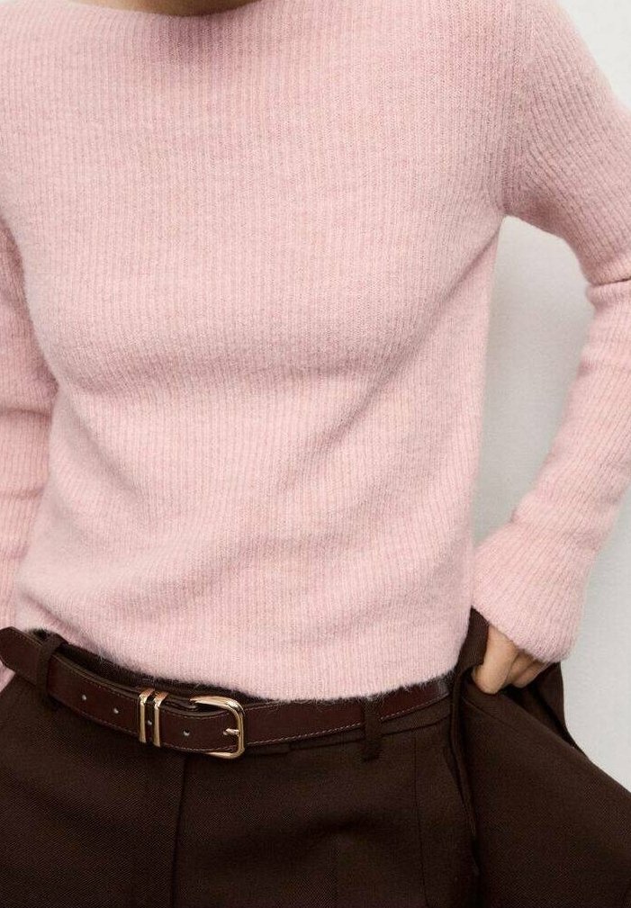 Personne portant un pull côtelé rose pâle rentré dans un pantalon marron foncé avec une ceinture marron assortie, la main dans la poche.