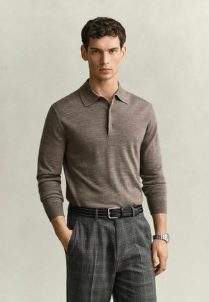 Jonge man met krullend haar, die een bruin poloshirt met lange mouwen draagt, grijze geruite broek, zwarte riem en zilveren horloge, staand met één hand in de zak.