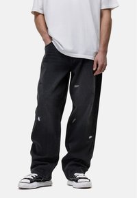 Jeans in denim scuro con una vestibilità ampia, caratterizzati da toppe scolorite e dettagli sfilacciati. Abbinati a sneakers bianche e nere e a una T-shirt bianca.