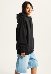 Schwarzer, übergroßer Hoodie mit Kängurutasche und Kordelzügen, kombiniert mit hellblauen Jeansshorts. Hat eine lockere Passform und eine weiche Textur.