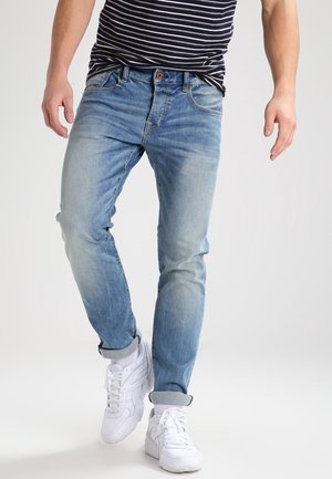 Mann trägt hellblaue hochgekrempelte Jeans, gestreiftes marineblau-weißes Hemd und weiße Turnschuhe, steht mit einem Bein leicht nach vorne.