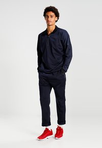 Pullover blu navy a maniche lunghe con zip a un quarto, abbinato a pantaloni blu navy a sigaretta e sneakers rosse. Tessuto liscio, vestibilità atletica.
