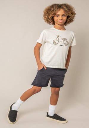 Kind met krullend haar, gekleed in een wit T-shirt met een scooterafbeelding en marineblauwe korte broek, staand met handen in de zakken tegen een beige achtergrond.