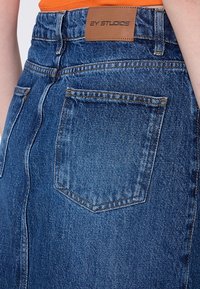 Gros plan de l'arrière d'une jupe en denim bleu avec deux poches et un patch en cuir marron étiqueté "2Y STUDIOS" à la ceinture.