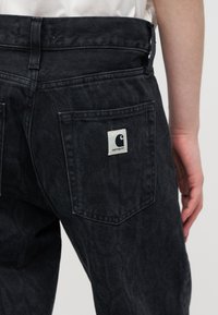 Σκούρα γκρι τζιν από denim με ένα λευκό μαξιλαράκι λογότυπου Carhartt στην πίσω τσέπη, φορεμένα από ένα άτομο με λευκό πουκάμισο σε ανοιχτό φόντο.