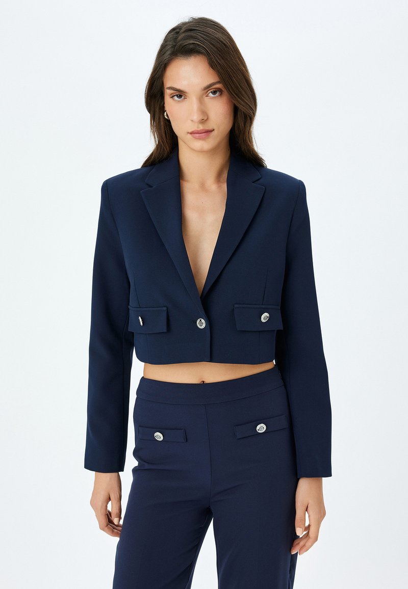 Navy cropped blazer dengan leher V yang dalam, kantong depan, dan kancing perak. Kain halus dan desain terstruktur untuk siluet yang pas.